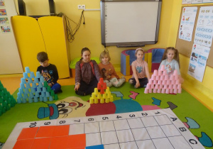 Dzieci siedzą przed ułożonymi z kubeczków kolorowymi wieżami.